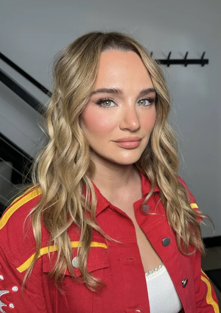 A young woman with wavy blonde hair and light makeup, wearing a red jacket with yellow details over a white top, standing indoors with a neutral background.