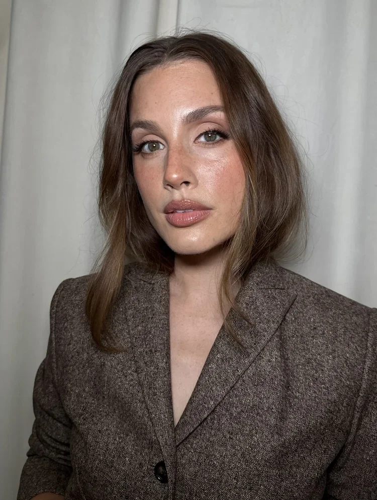 A woman with shoulder-length brown hair, wearing a brown blazer, looking at the camera with a neutral expression. Background consists of light-colored curtains.