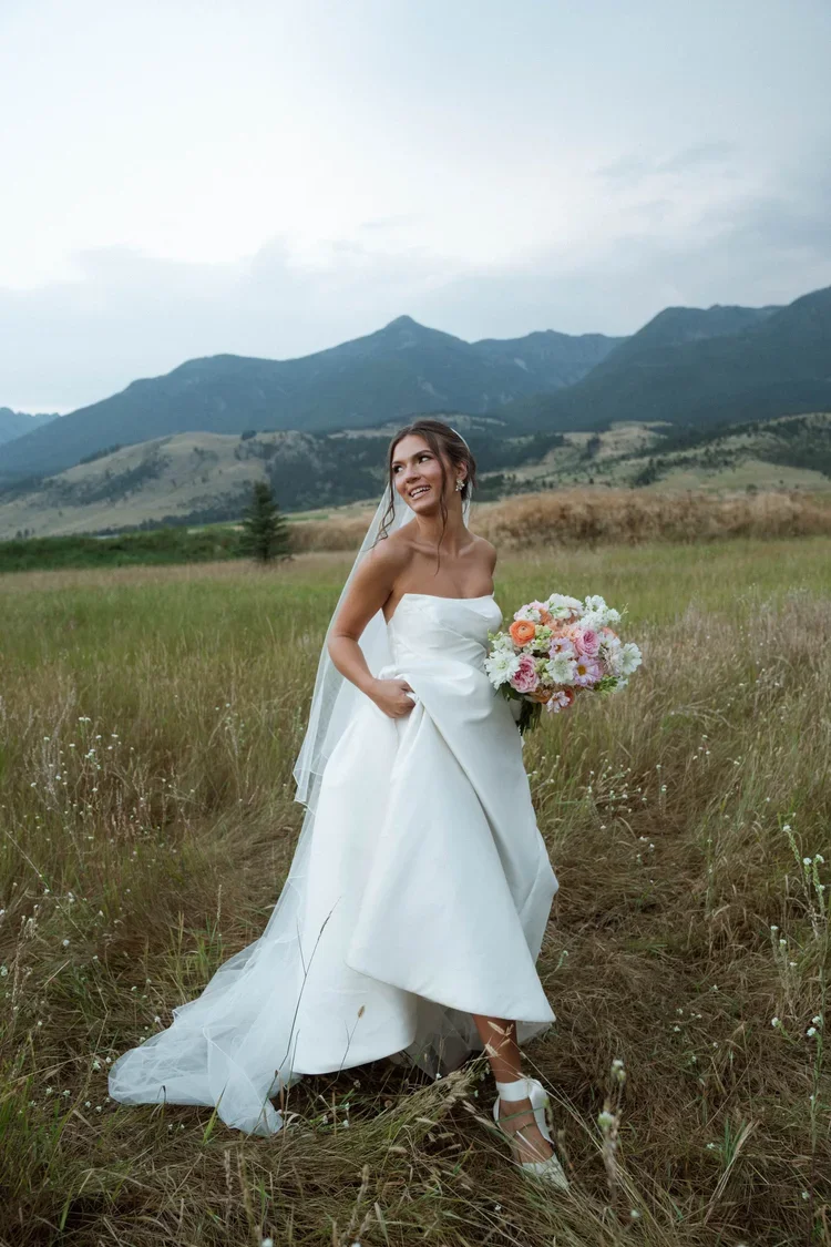 A bride in a white strapless wedding gown and veil holding a bouquet of pink, white, and peach flowers, standing in a grassy field with mountains in the background.