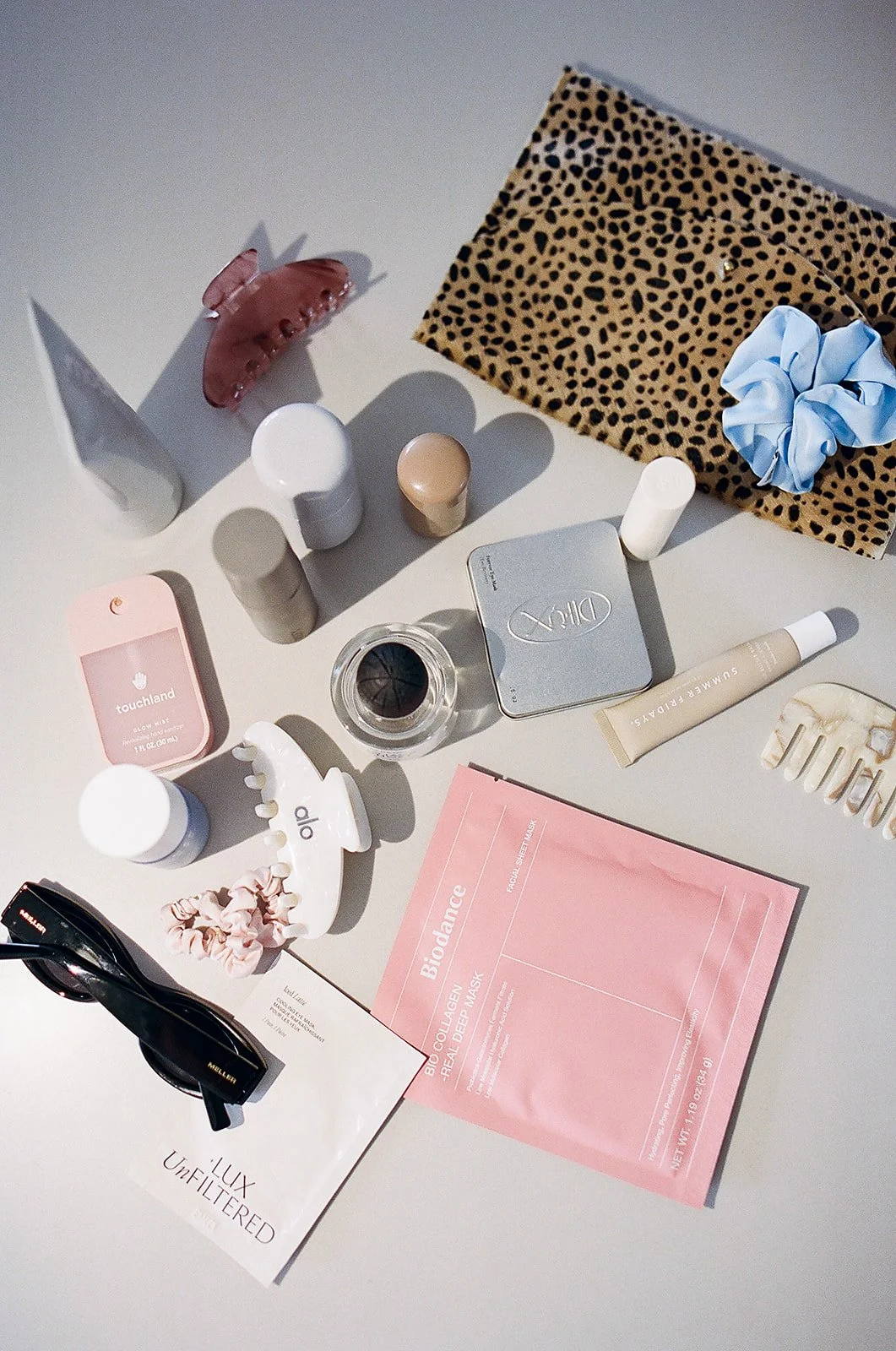 Flat lay of skincare, beauty, and fashion items on a gray surface, including a leopard print cloth, a blue scrunchie, skincare products, sunglasses, a pink facial mask packet, hair clips, and a small pink makeup sponge.