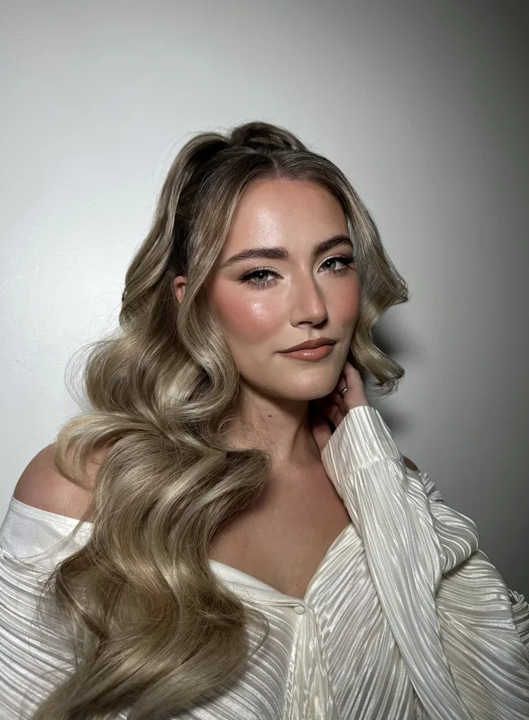 A woman with long, wavy blonde hair, wearing a white off-shoulder top, posing against a plain gray background.