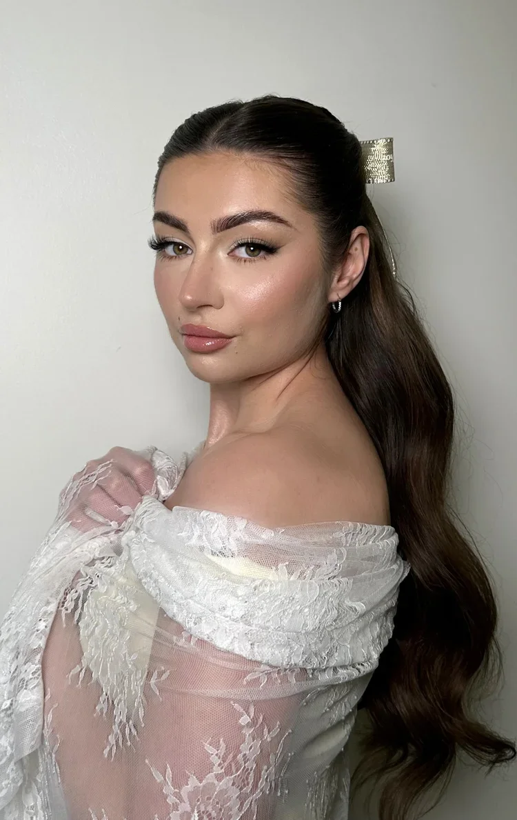 Young woman with long dark hair, wearing makeup, in a white lace off-shoulder top, posing against a plain wall.