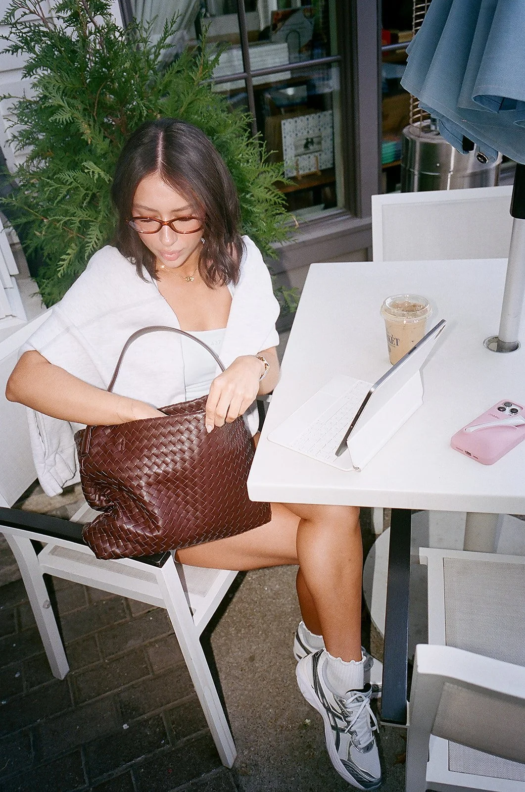 A woman sitting at an outdoor cafe table, wearing glasses, a white top, sneakers, and a watch, with a large woven brown purse on her lap. She has a drink, a tablet, and a pink phone on the table.