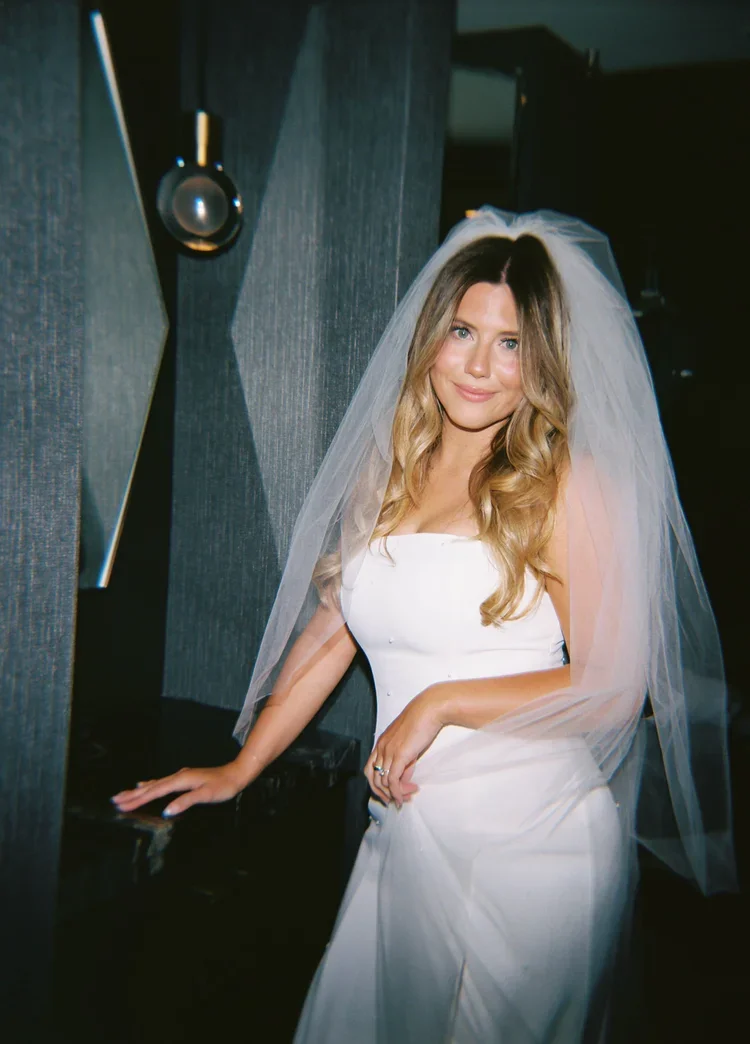 Woman in wedding dress with veil standing indoors, leaning on a surface, smiling.