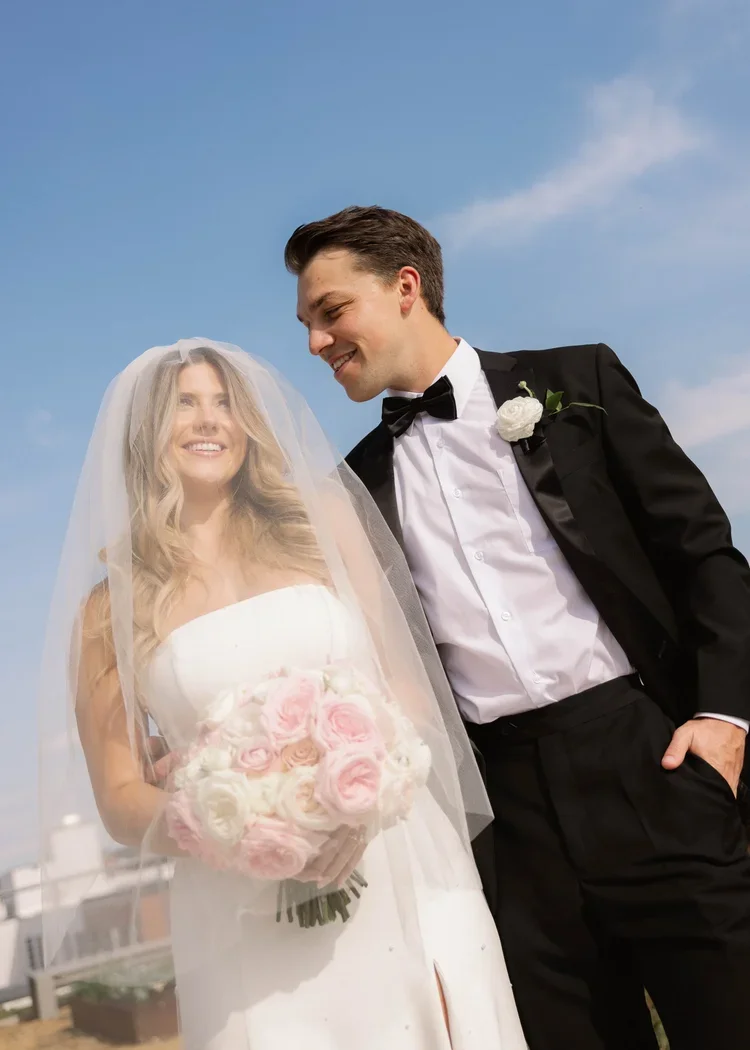 A bride and groom smiling together outdoors on their wedding day, with a blue sky and clouds in the background. The bride is holding a bouquet of pink and white roses, wearing a white wedding dress and veil. The groom is in a black tuxedo with a whit