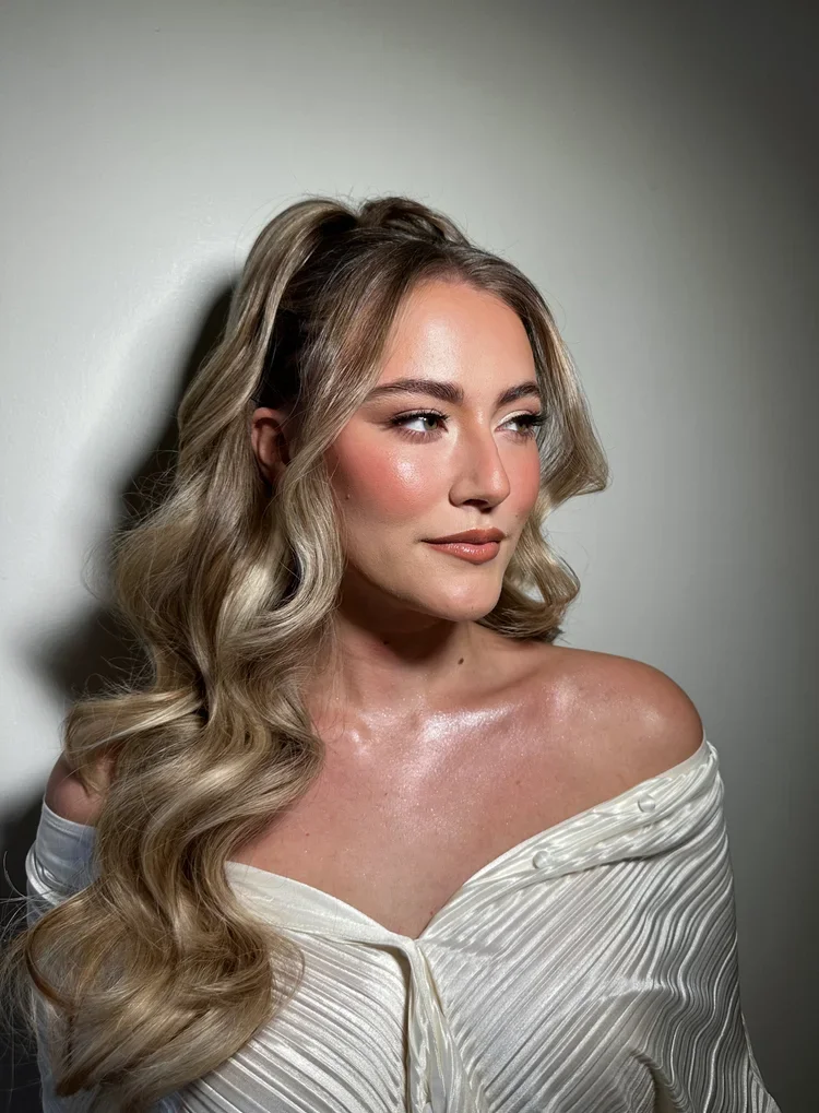 A portrait of a woman with styled blonde hair, wearing an off-the-shoulder white top, against a plain light-colored background.