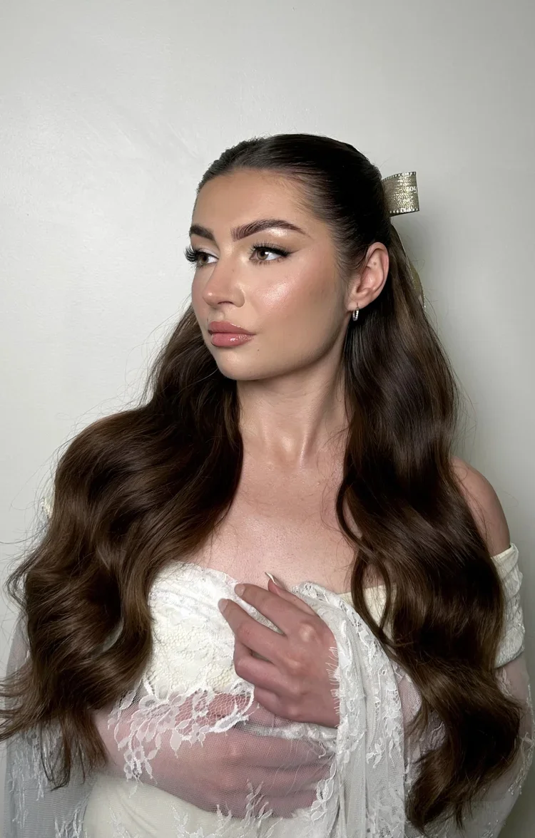 A young woman with long, wavy brown hair, styled with a gold ribbon hair accessory, wearing a white lace off-the-shoulder dress, posing against a plain white wall.