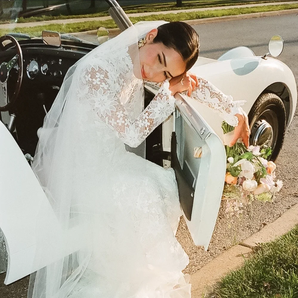 A bride in a white lace wedding dress and veil sitting in an open vintage car with a bouquet of flowers placed nearby.