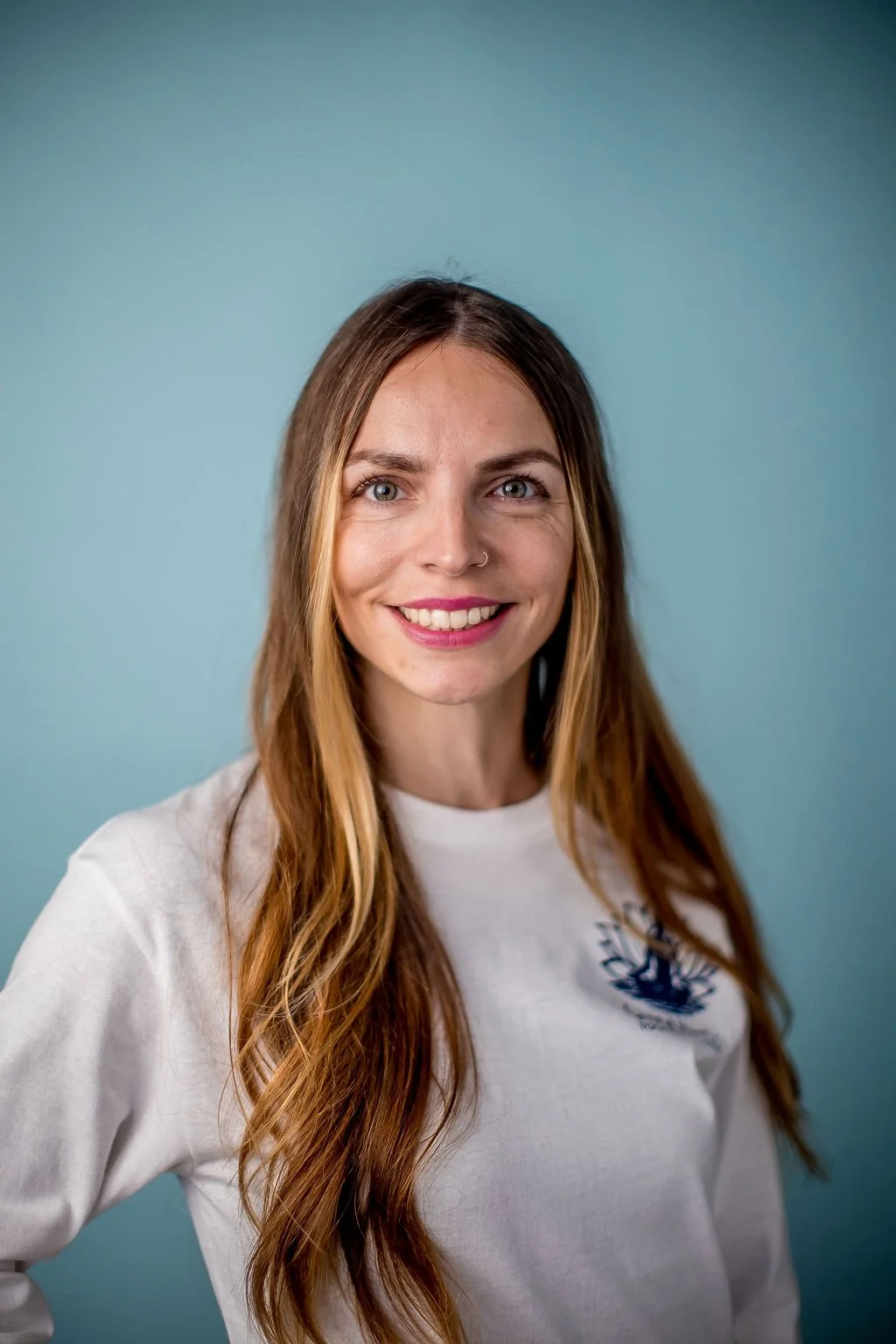 Portrait of a smiling young woman with long, wavy red hair and a nose ring, wearing a white shirt, standing in front of a light blue background.