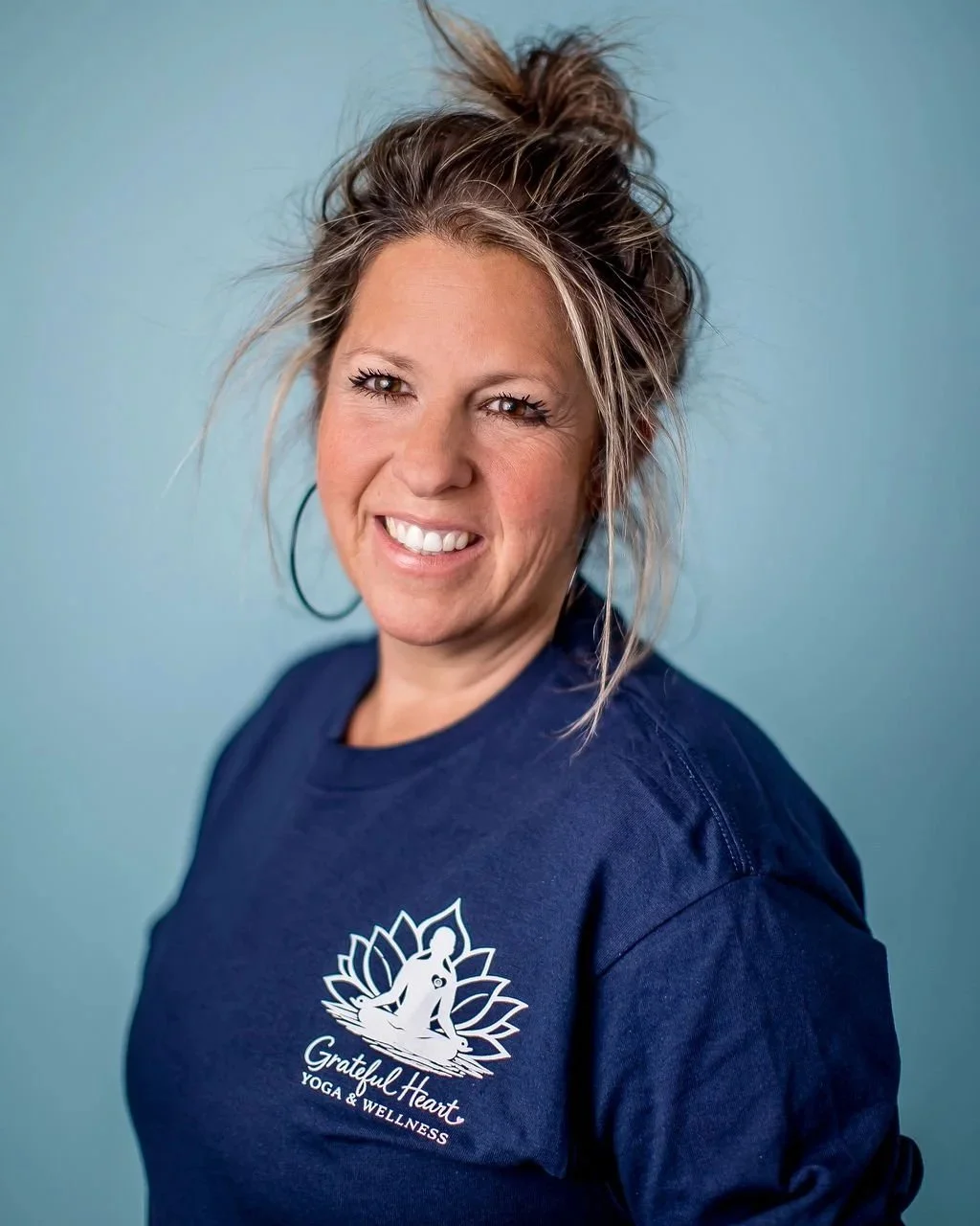 A woman with light brown hair styled in a messy bun, smiling, wearing a navy blue shirt with a logo for 'Grateful Heart Yoga & Wellness' on it, standing against a light blue background.