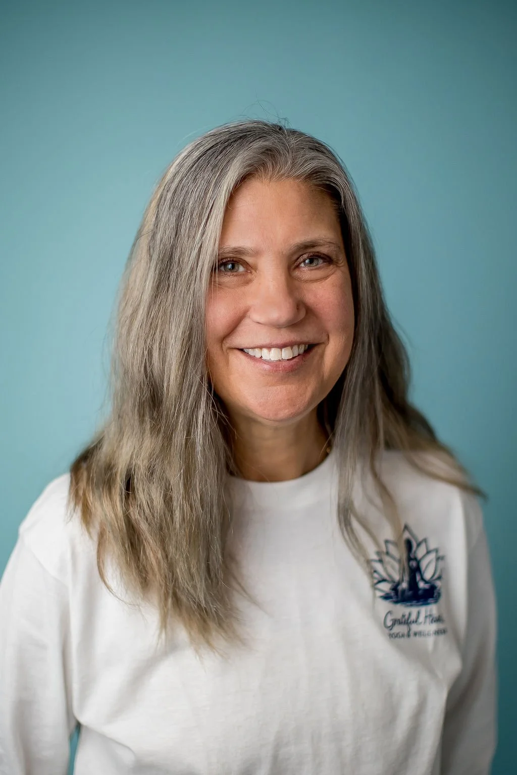 A woman with long gray hair smiling, wearing a white shirt with a logo on it, standing against a blue background.