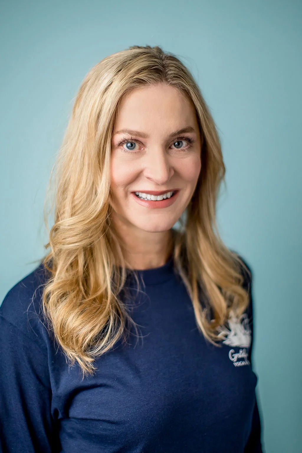 Portrait of a smiling woman with long wavy blonde hair, wearing a navy blue shirt, against a light blue background.