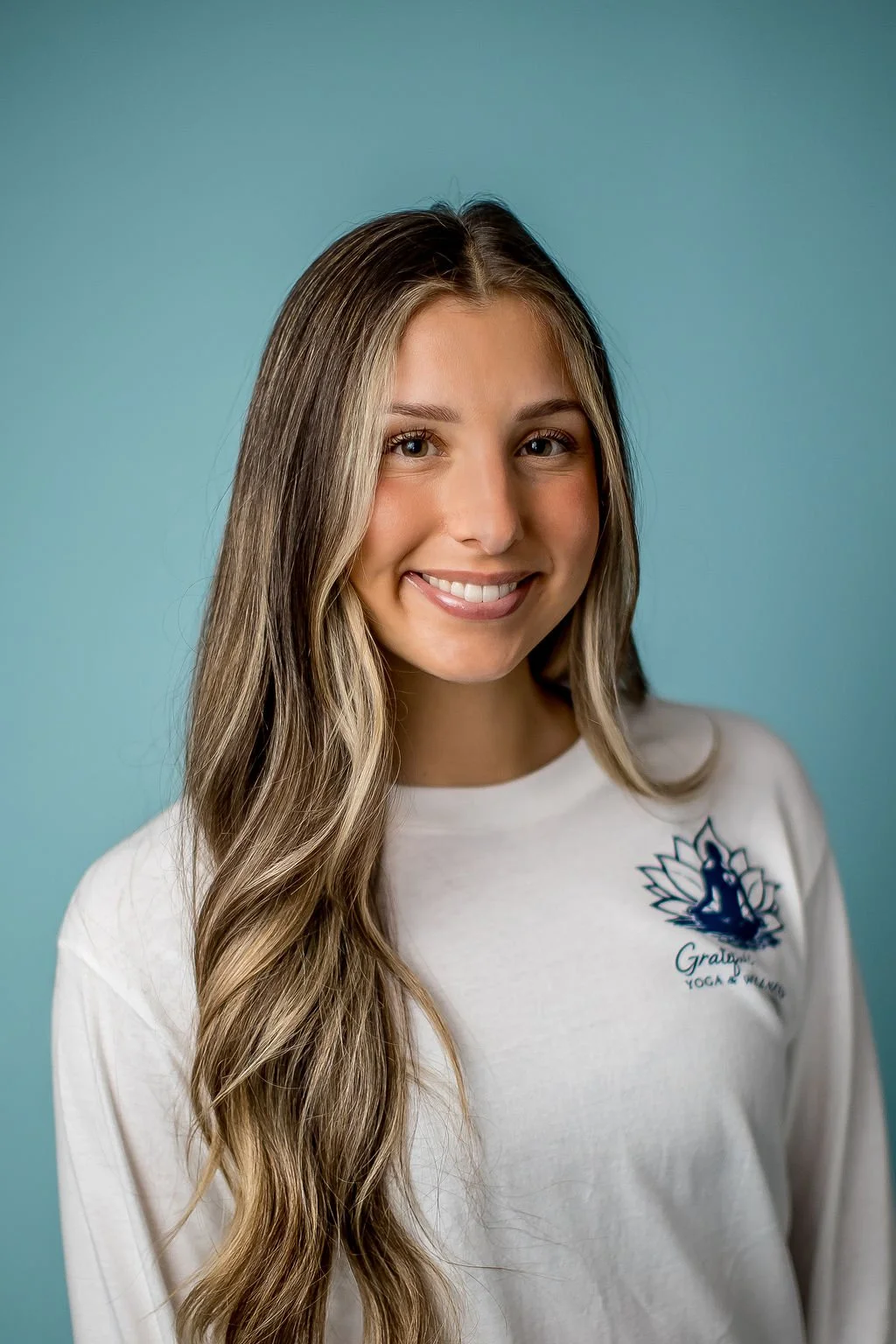 A young woman with long, wavy blonde hair smiling, wearing a white shirt with a yoga logo, against a light blue background.