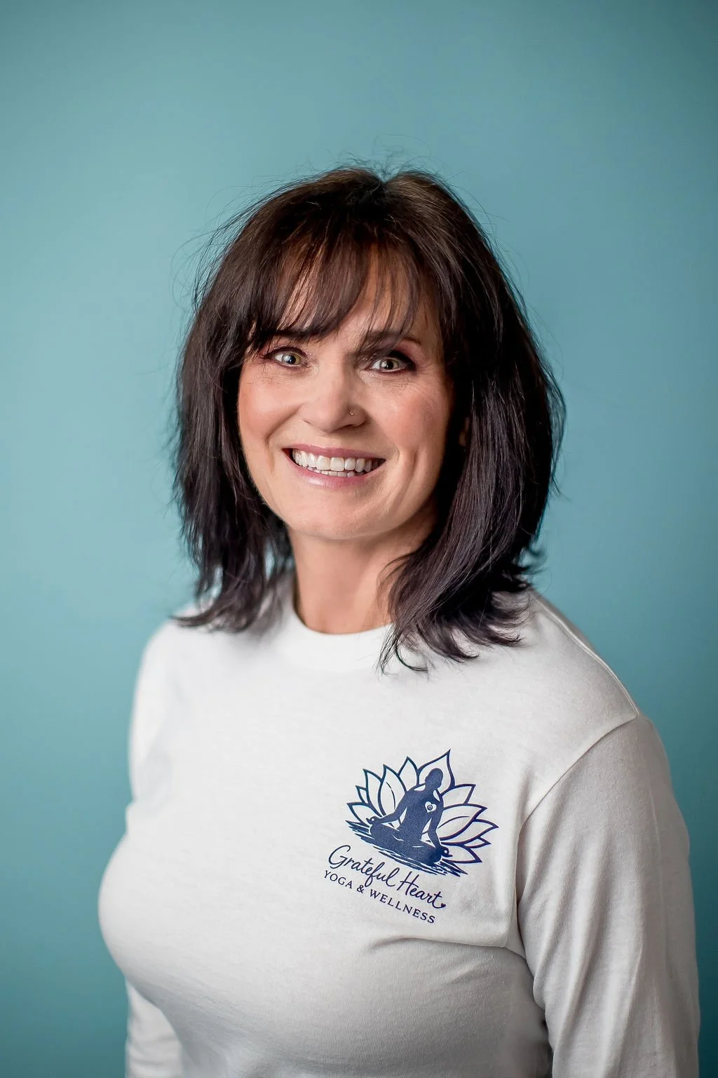 A smiling woman with shoulder-length dark brown hair, wearing a white shirt with a logo that reads "Grateful Heart Yoga & Wellness" and a lotus flower design, standing against a teal background.
