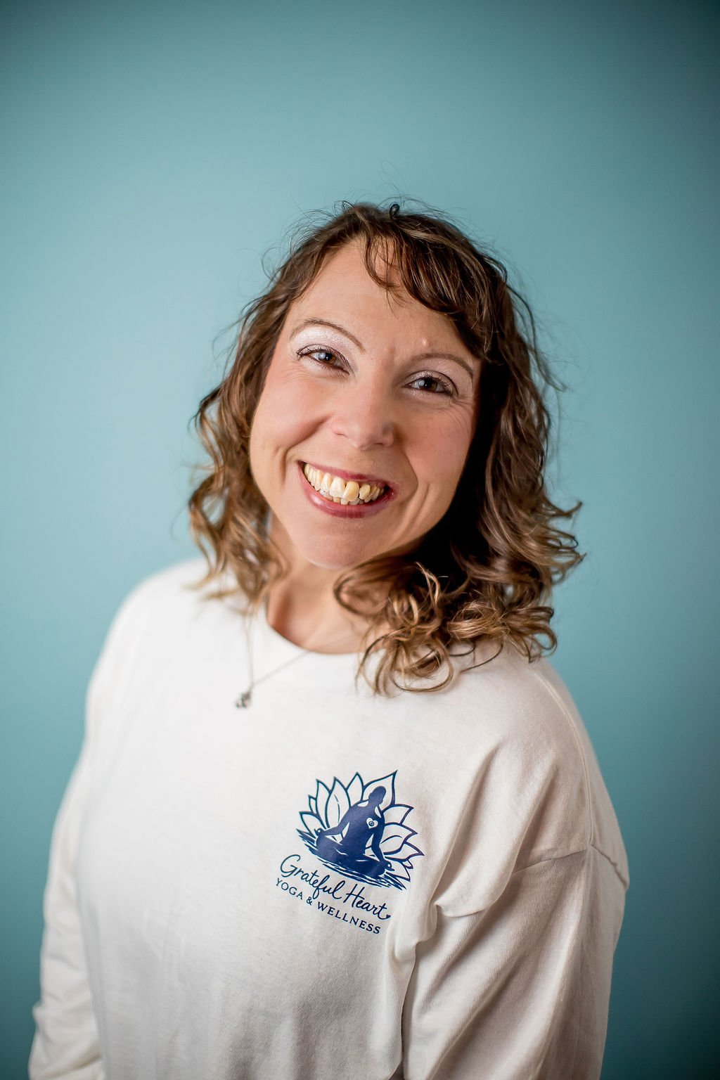 A woman smiling, wearing a white shirt with a logo for "Grateful Heart Yoga & Wellness" on it, standing against a light blue background.