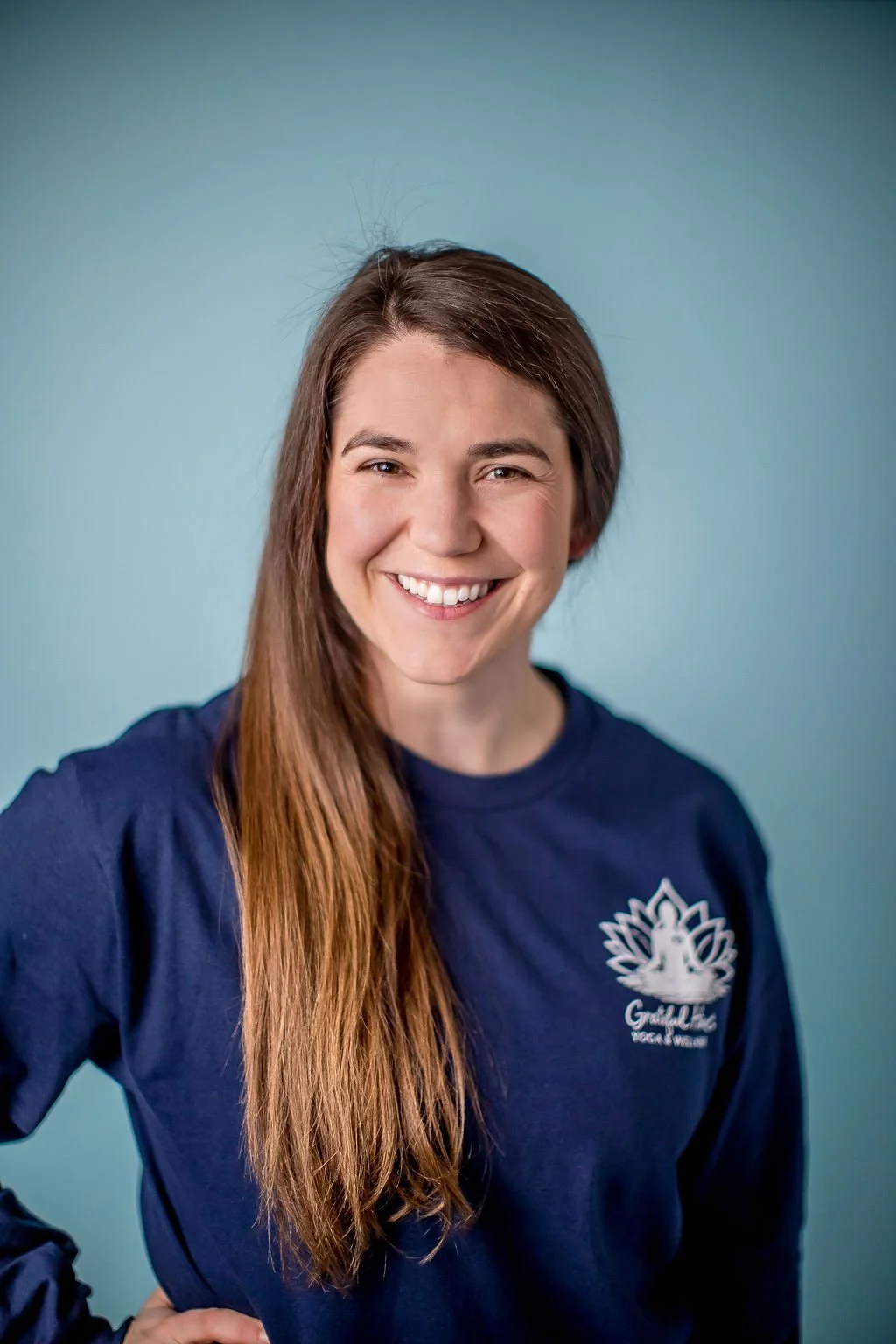 Portrait of a smiling young woman with long brown hair, wearing a navy blue shirt with a lotus flower logo, against a plain light blue background.
