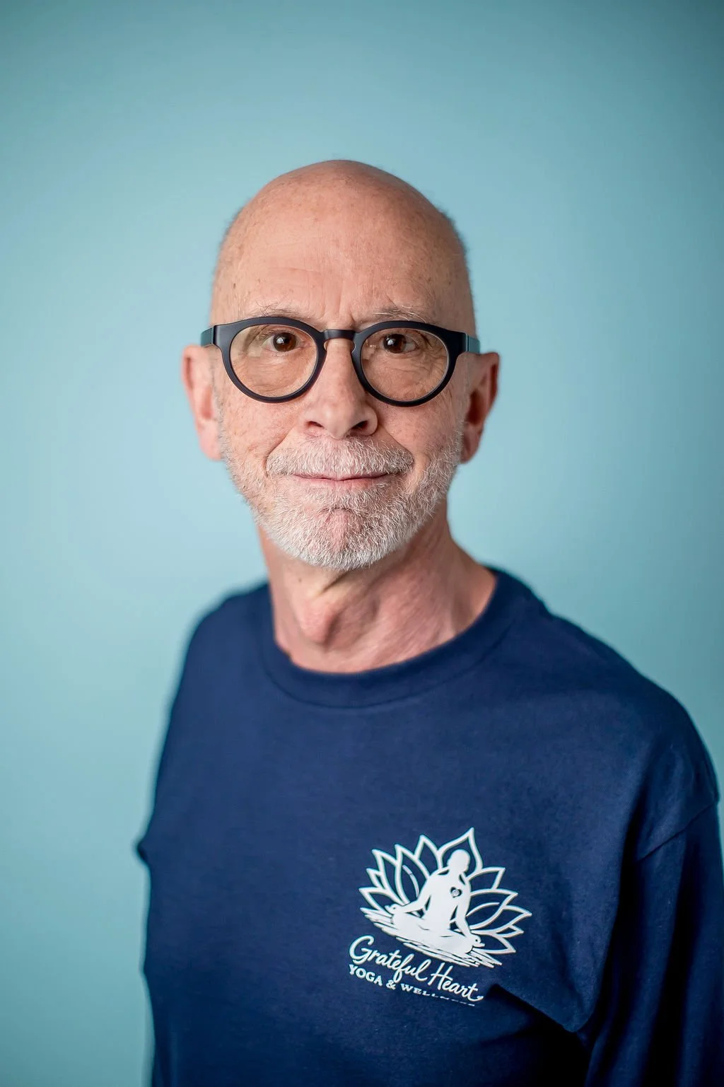 A smiling older man with a bald head, gray beard, and round glasses, wearing a navy blue T-shirt with a yoga logo and the words 'Grateful Heart Yoga & Wellness' against a light blue background.