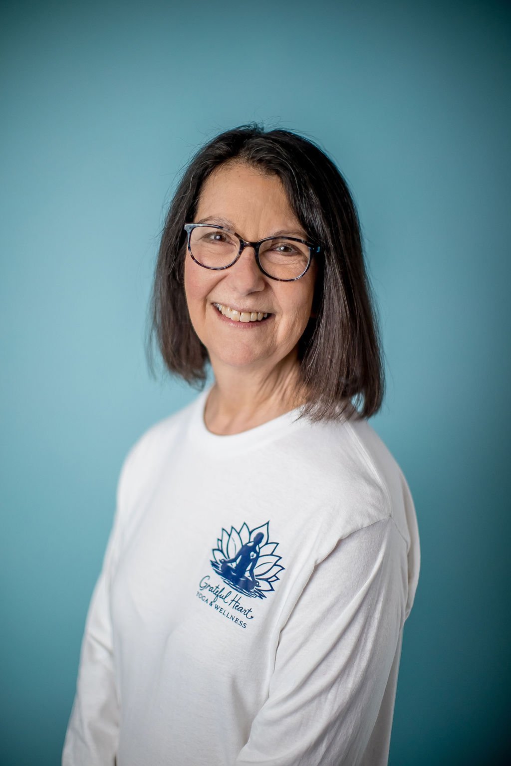 A smiling woman with shoulder-length dark hair, glasses, and a white shirt featuring a logo with a lotus flower and the text "Grateful Heart Yoga & Wellness" against a solid blue background.