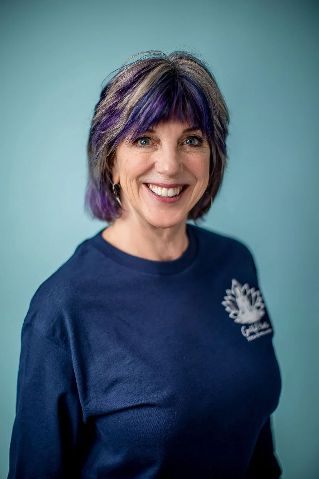 A woman with shoulder-length, multi-colored hair including purple and blonde, smiling against a plain light blue background, wearing a navy blue shirt with a logo on the chest.