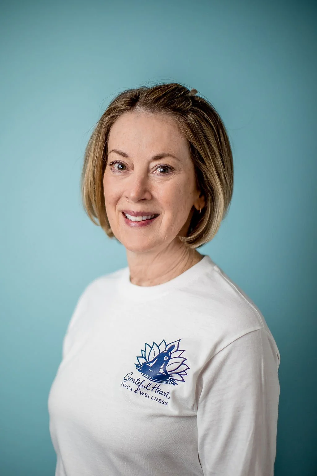 A woman with short blonde hair smiling, wearing a white T-shirt with a logo that says 'Grateful Heart Yoga & Wellness', standing against a light blue background.