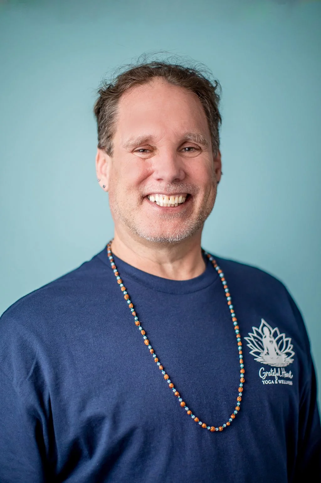 A man with short brown hair, a beard, and earrings, smiling at the camera. He is wearing a navy blue shirt with a logo that says 'Grateful Heart Yoga & Wellness' and a beaded necklace, standing against a light blue background.