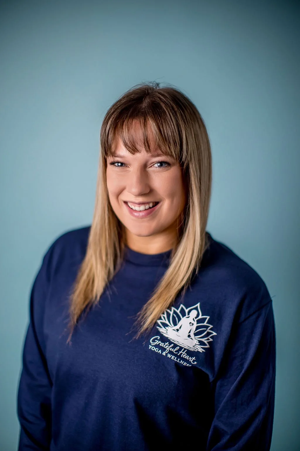 Smiling woman with light brown hair and bangs wearing a navy blue shirt with a yoga wellness logo.