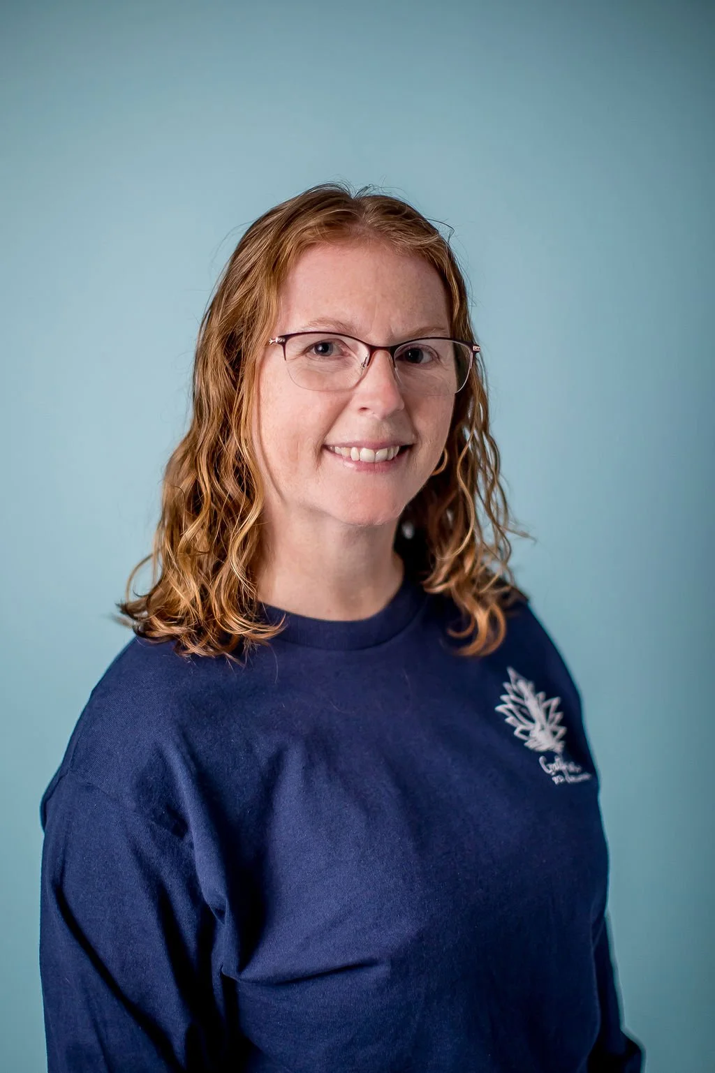 A woman with shoulder-length curly red hair and glasses smiling in front of a light blue background.