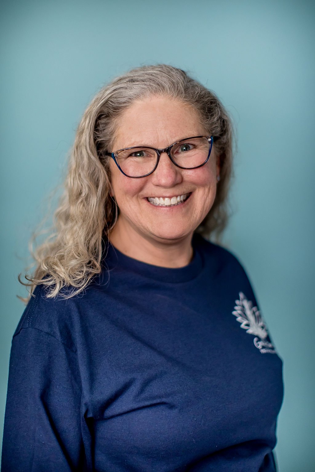 A woman with curly gray hair, glasses, and a blue shirt is smiling against a light blue background.