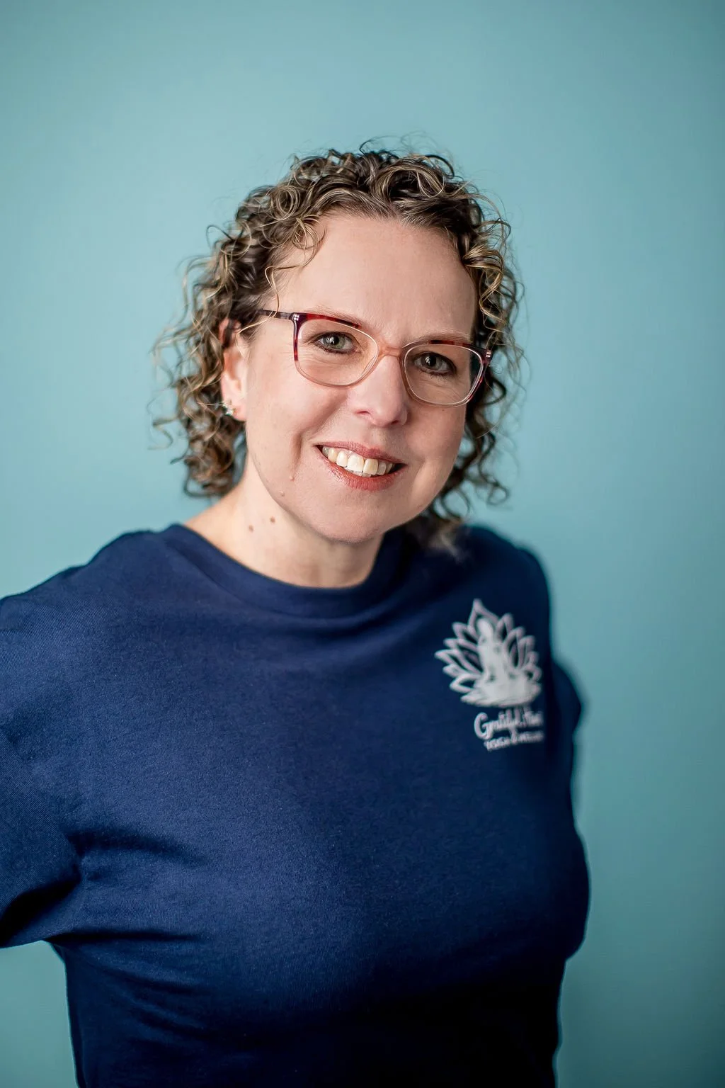 A woman with curly hair and glasses, smiling, wearing a navy blue shirt with a logo, standing against a light blue background.