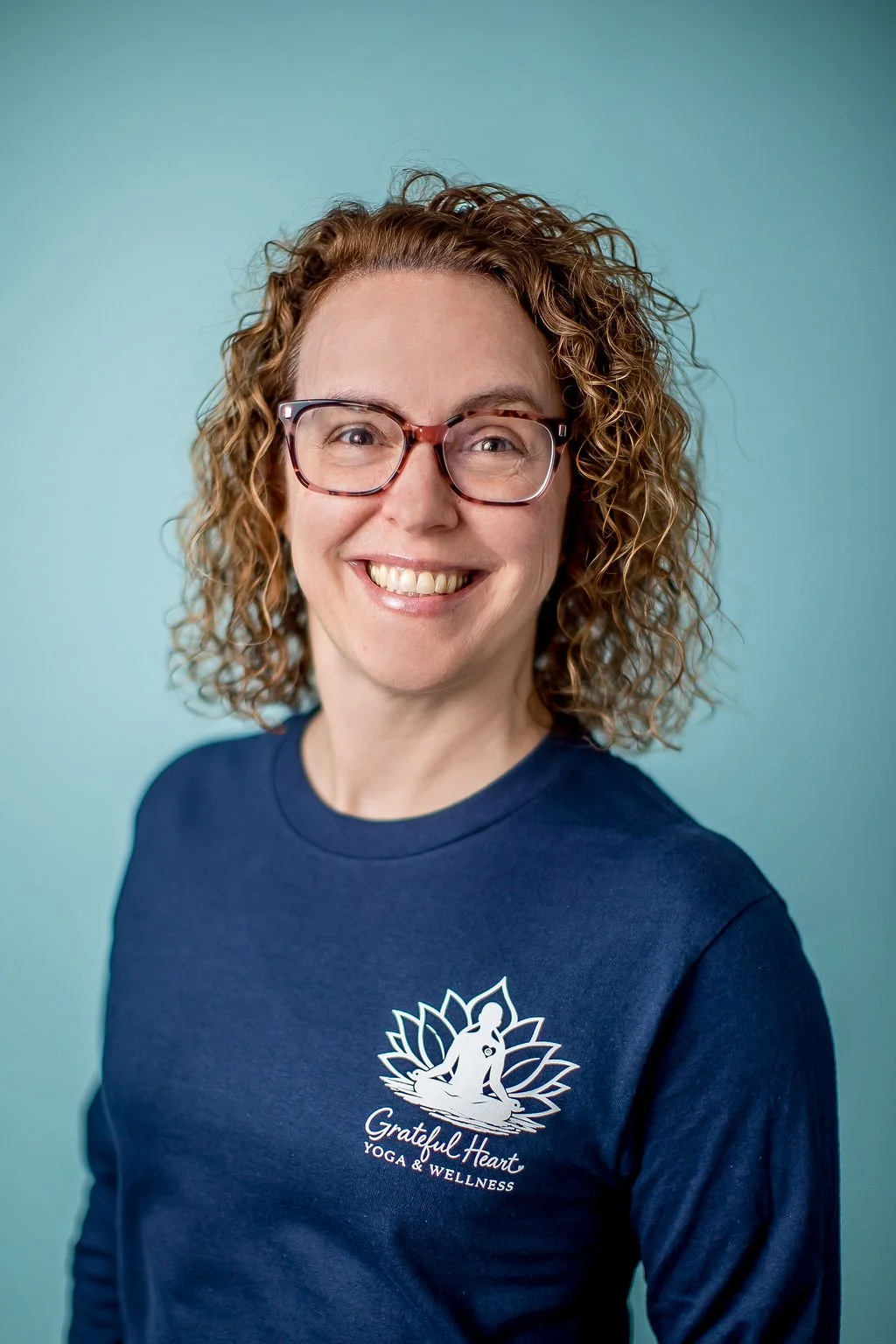 Portrait of a woman with curly hair, glasses, smiling, wearing a navy blue shirt with 'Grateful Heart Yoga & Wellness' logo, against a light blue background.