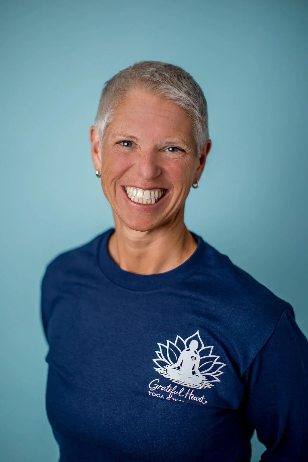 Smiling woman with short gray hair wearing a dark blue T-shirt with a yoga logo and text against a light teal background.