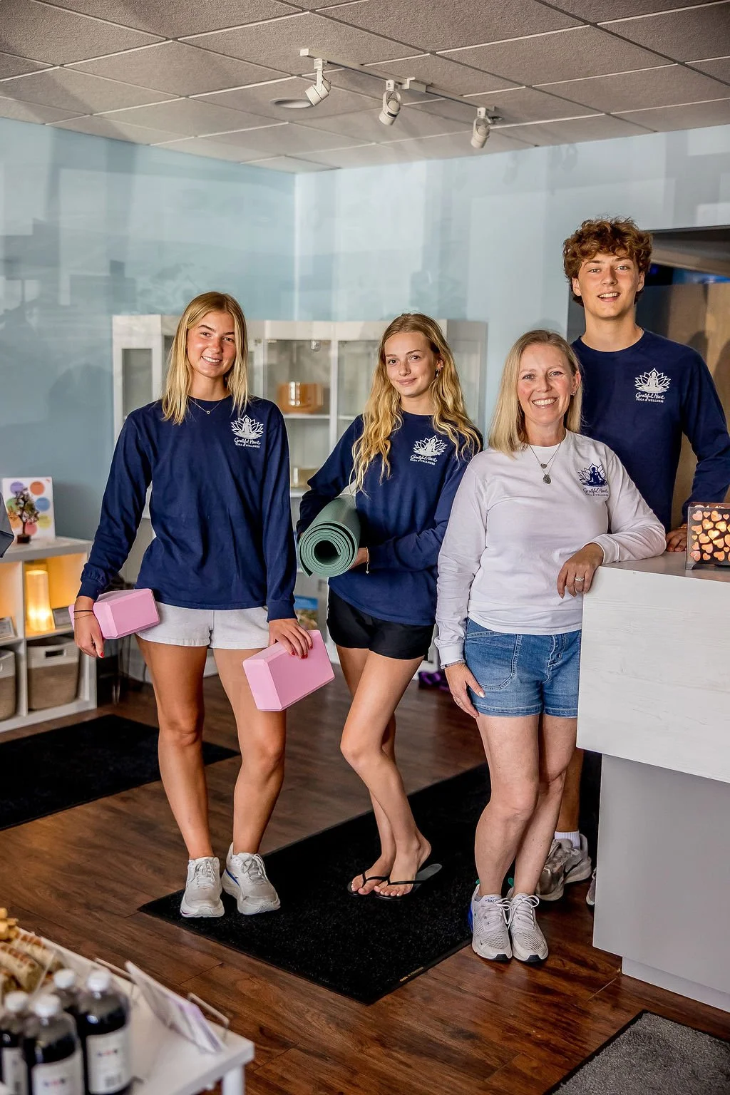 Four smiling women standing together inside a wellness or yoga studio, some holding pink yoga blocks, with a reception desk visible in the background.