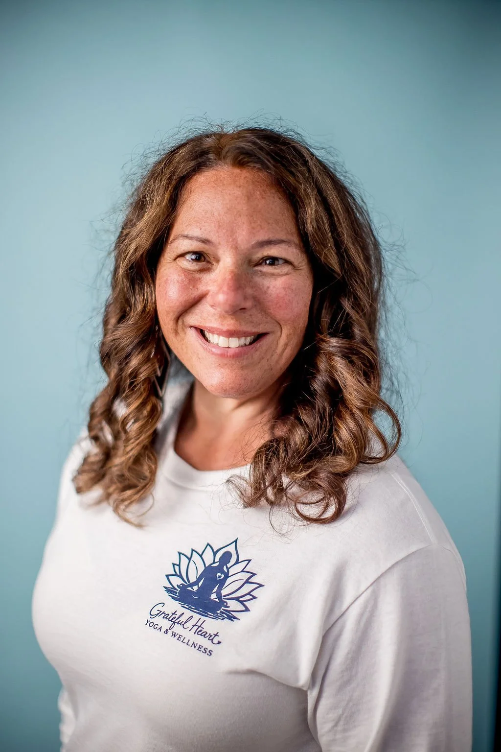 A smiling woman with curly brown hair wearing a white t-shirt with a logo that says 'Grateful Heart Yoga & Wellness' and a graphic of a person meditating with a lotus flower background, against a blue background.