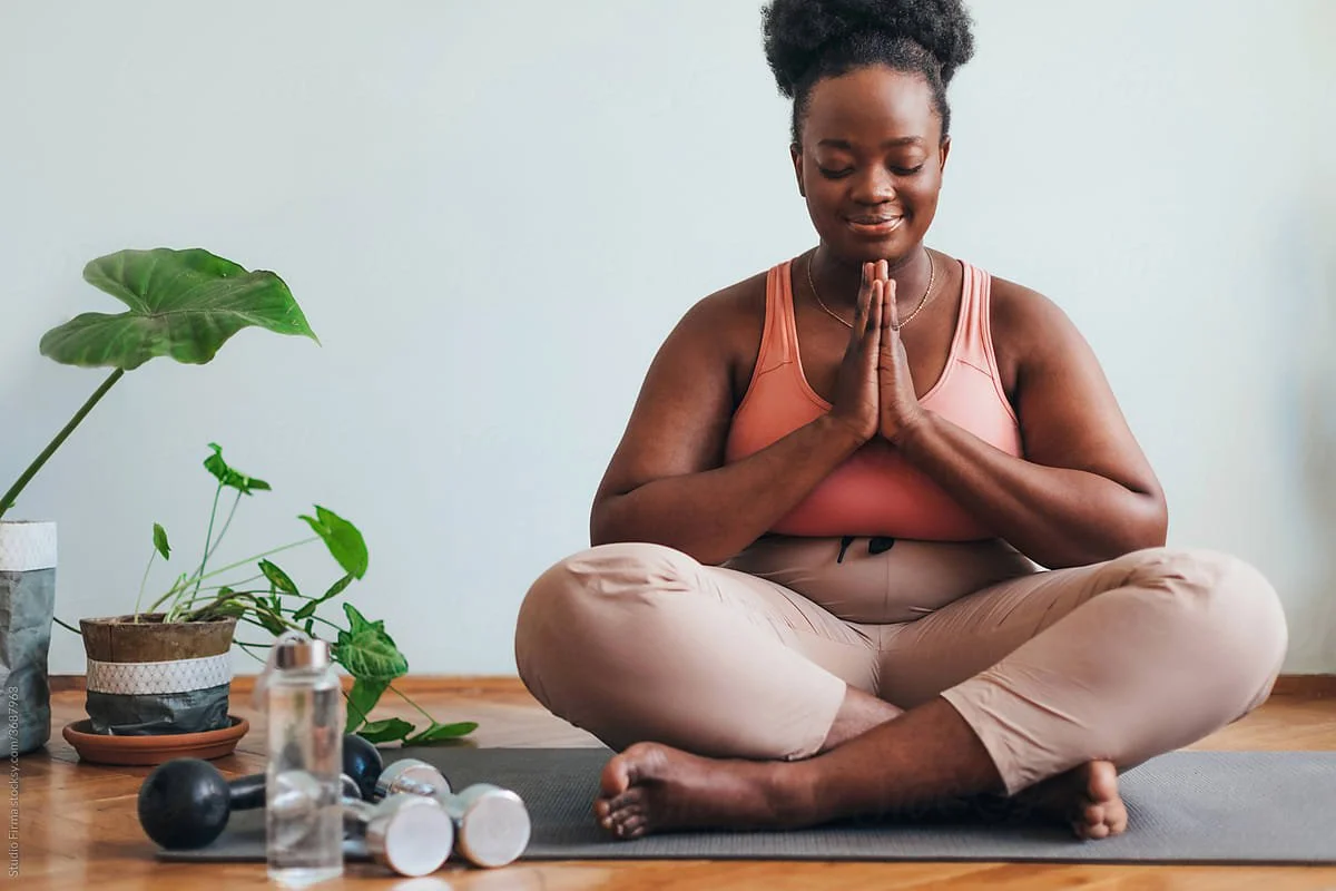 A woman with natural curly hair in a pink sports top and beige pants sitting cross-legged on a yoga mat with her hands in prayer position, meditating indoors next to potted plants and workout equipment.