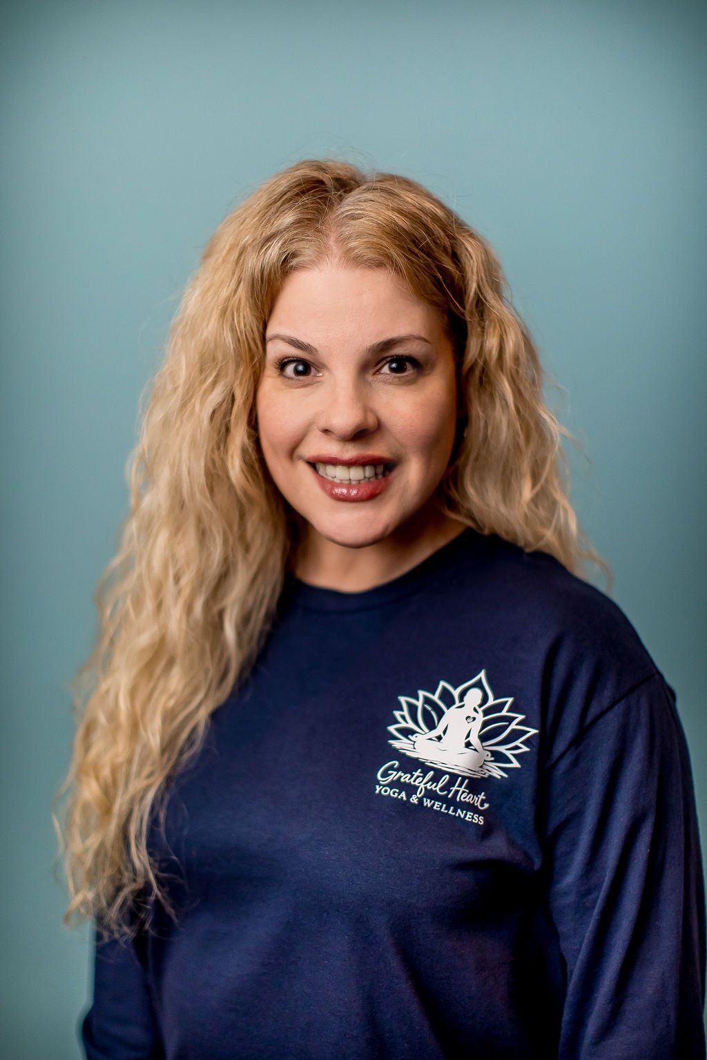 A woman with long, curly blonde hair smiling at the camera, wearing a navy blue T-shirt with a logo that reads "Grateful Heart Yoga & Wellness" and features a sitting yoga pose and a lotus flower.