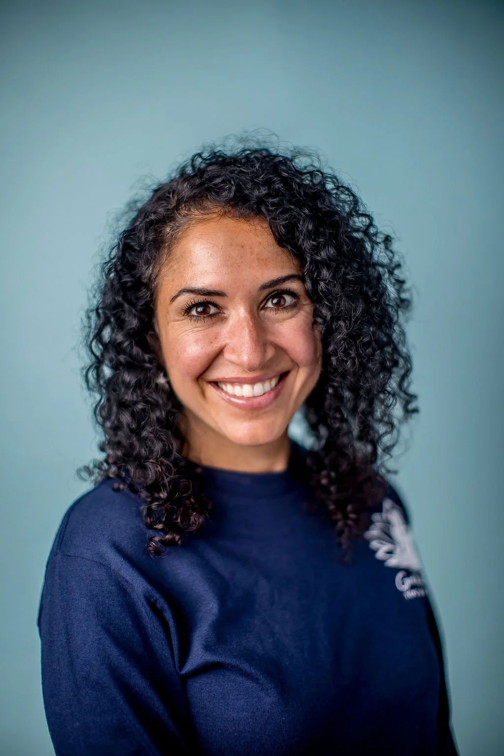A woman with curly dark hair smiling against a light blue background.