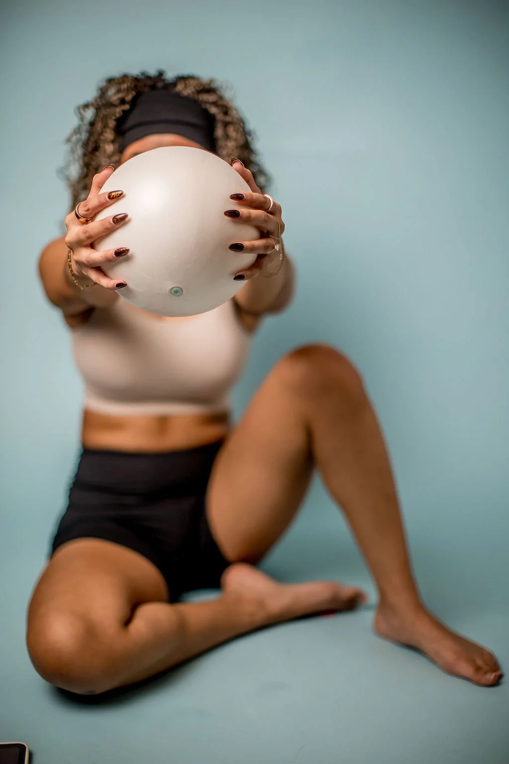 A woman sitting on the floor holding a round object, possibly a smart speaker or weather device, towards the camera. She has curly hair, a black headband, tattoos on her arms, and is wearing a beige sports bra and black shorts, with a light blue background.