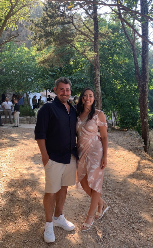 A man and woman stand together outdoors in a wooded area, smiling. The man wears a black shirt and beige shorts; the woman wears a pink dress with a pattern. Other people are visible in the background.