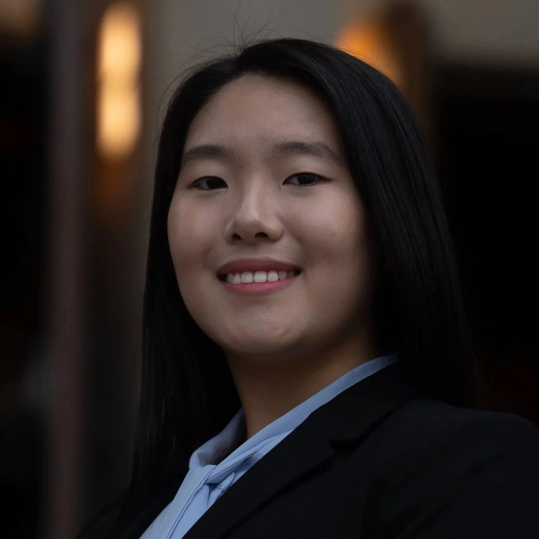 Close-up of a young woman smiling, wearing a black academic gown and cap, with a blurred background, possibly at graduation.