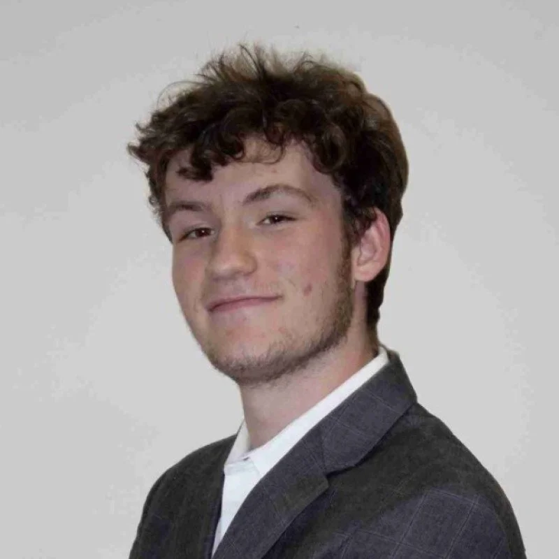 Young man with curly brown hair and slight beard, wearing a dark blazer and white shirt, smiling with a playful expression against a plain gray background.