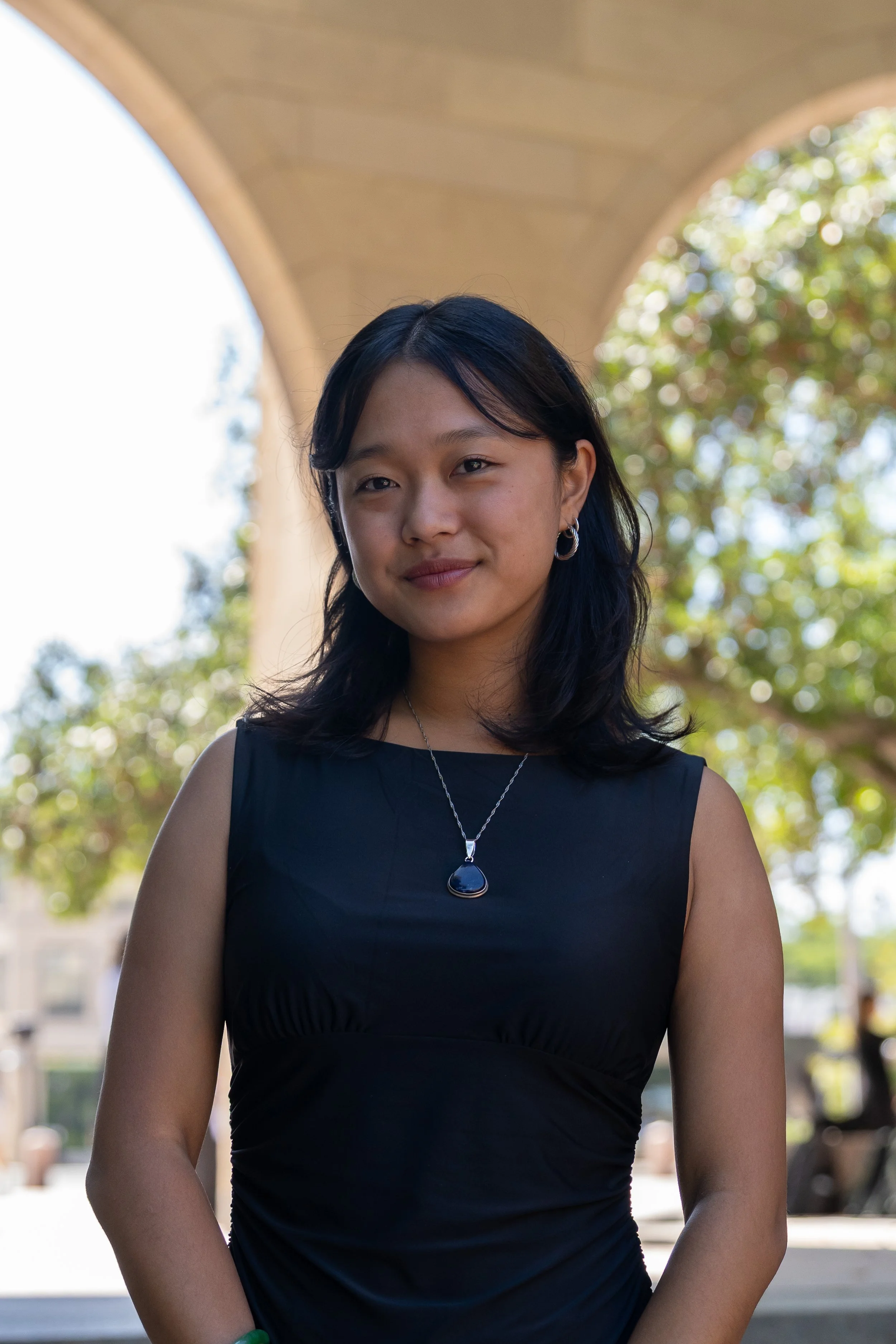 Yasmine Chang. A young woman with shoulder-length black hair and earrings, wearing a gray shirt and black blazer, standing outdoors with a blurred background of a bridge and greenery, smiling at the camera.