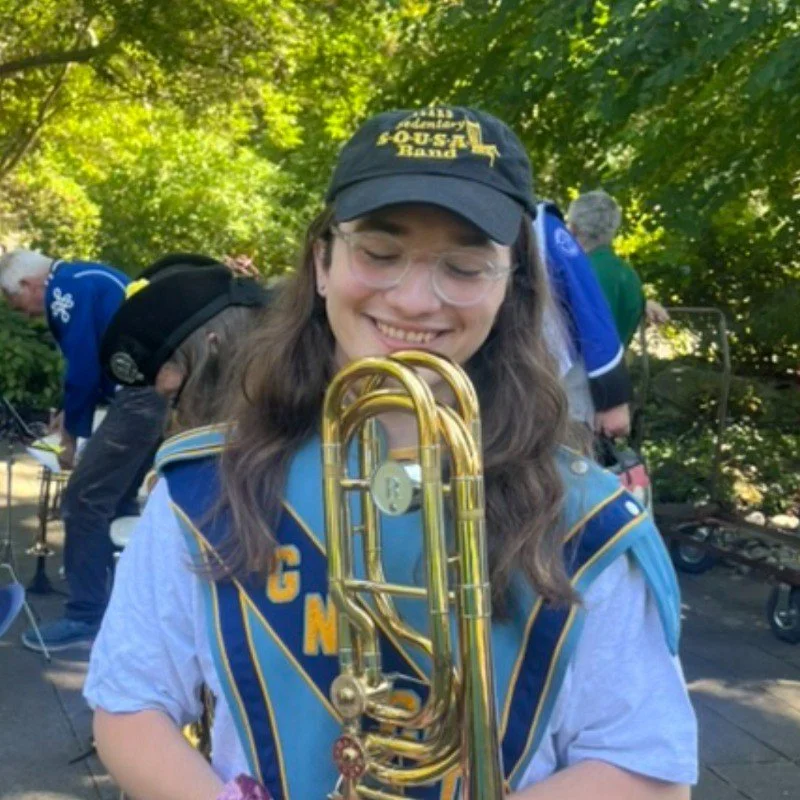 A young woman smiling and holding a trombone outdoors, wearing a band uniform and cap, with other band members in the background and lush green trees overhead.