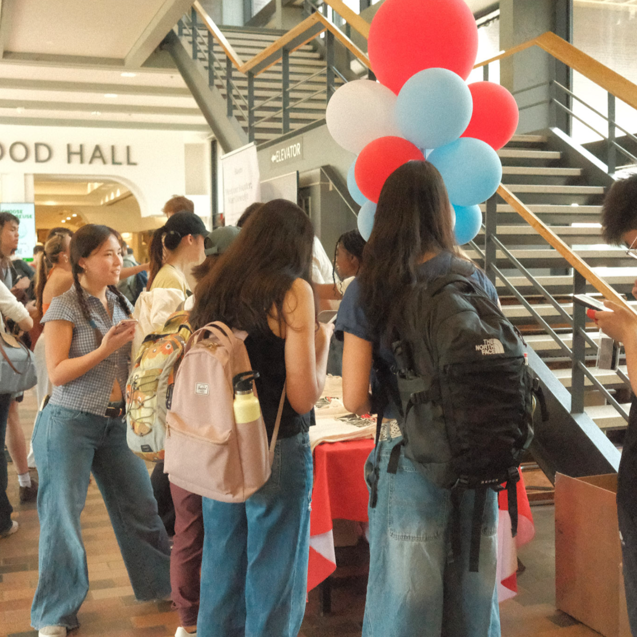 Group of young women at a registration or information table, with a balloon decoration in a busy indoor setting.