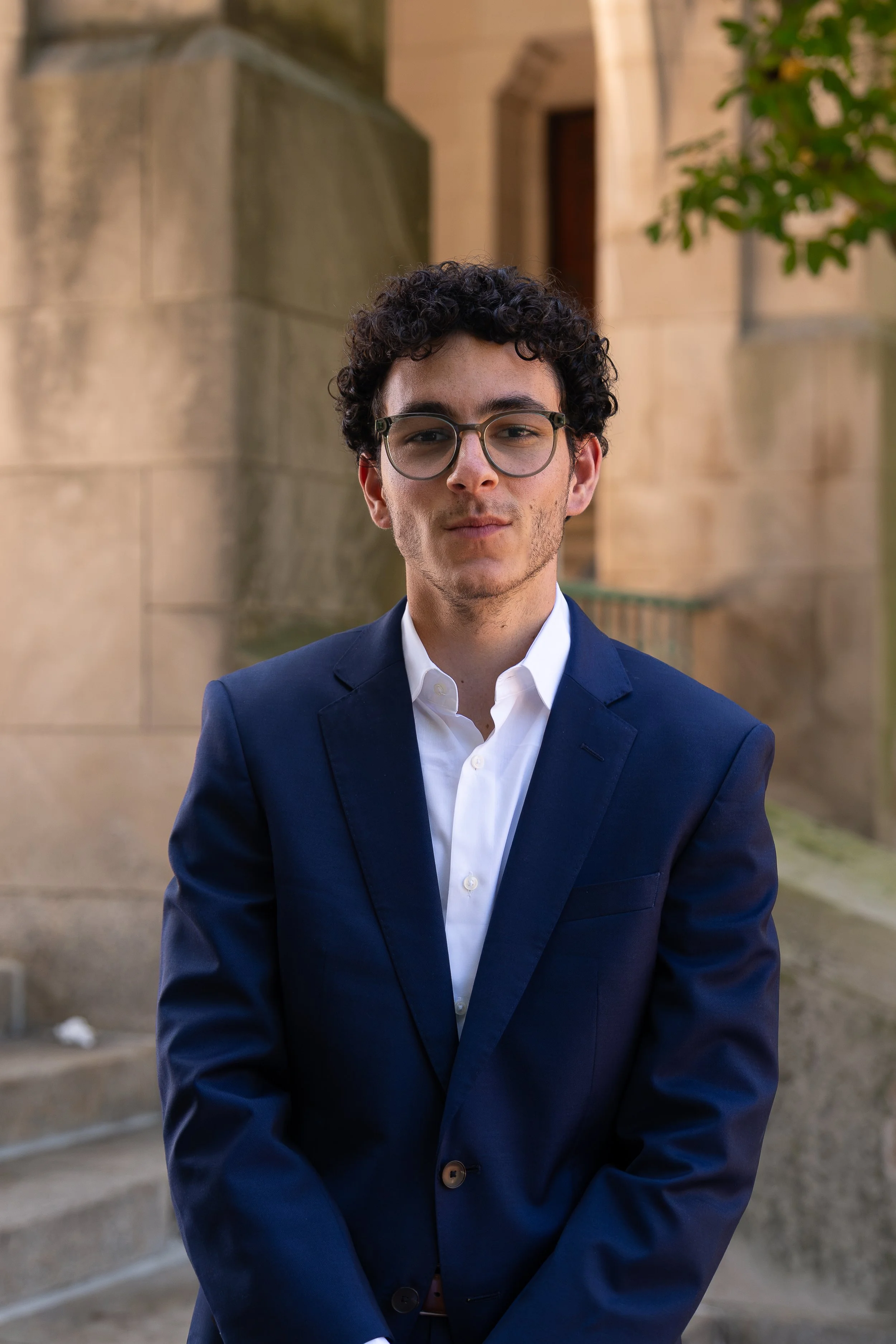 Young man in a navy blue suit and white shirt, smiling and wearing glasses, standing against a plain background.