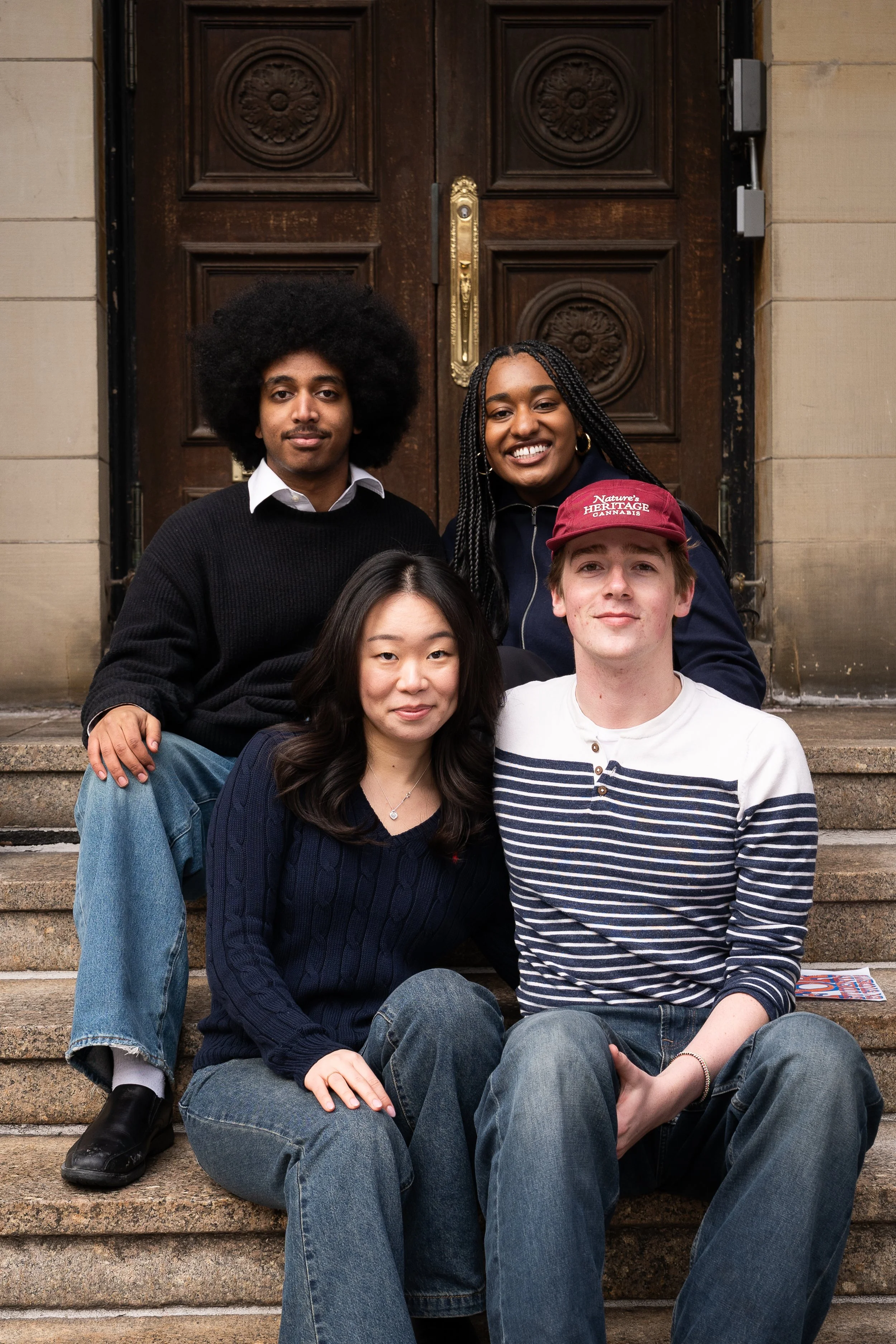 A group of five young adults sitting on the steps of a building with a large, ornate wooden door behind them. They are smiling and looking at the camera, dressed casually.
