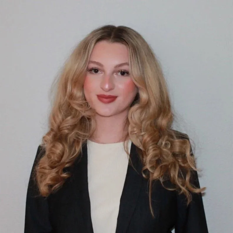 A young woman with long, curly blonde hair, wearing a black blazer over a white top, standing against a plain white wall.