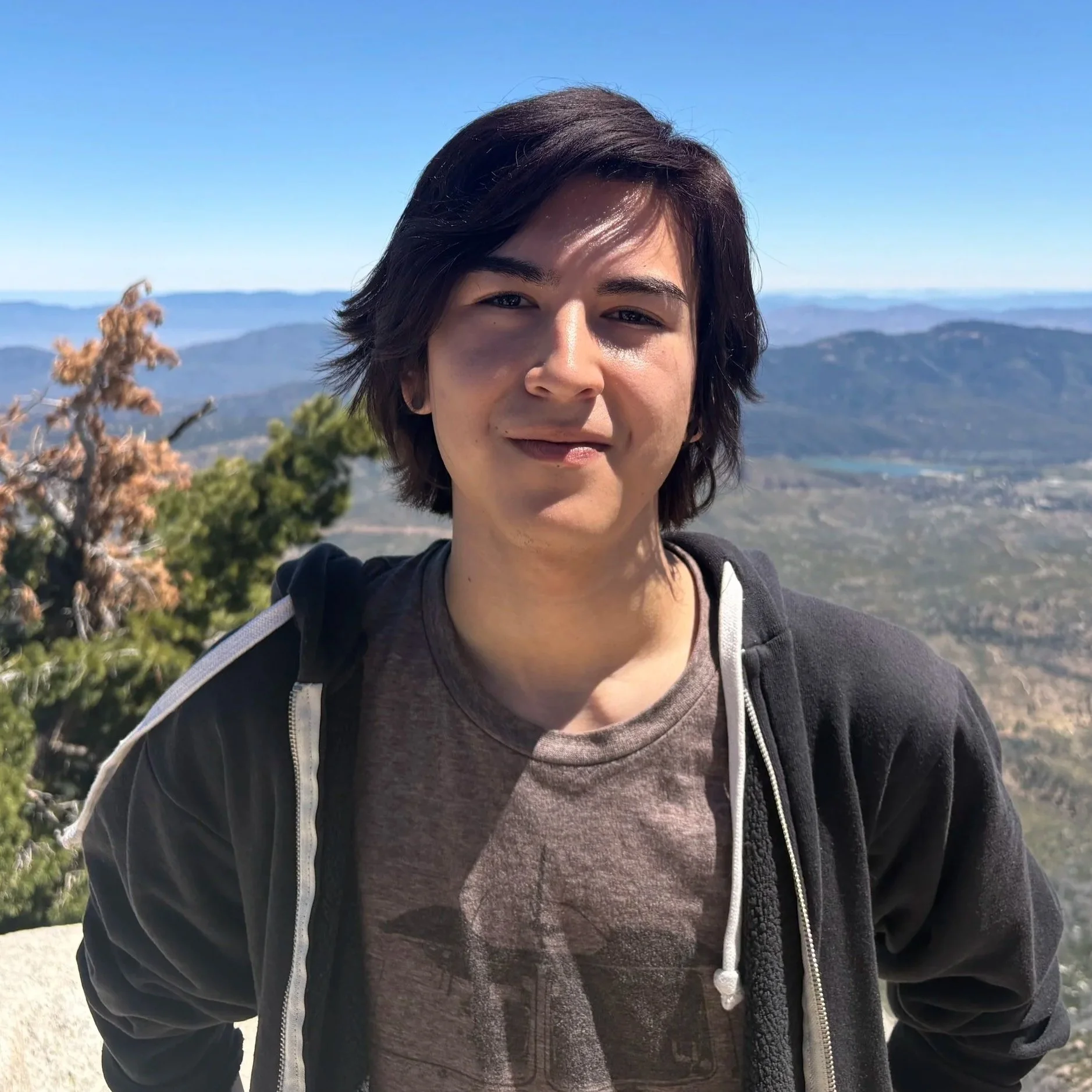 A person with short dark hair and light skin taking a selfie outdoors on a hillside with mountains and a clear blue sky in the background. The person is wearing a dark jacket with a zipper and a gray T-shirt underneath.