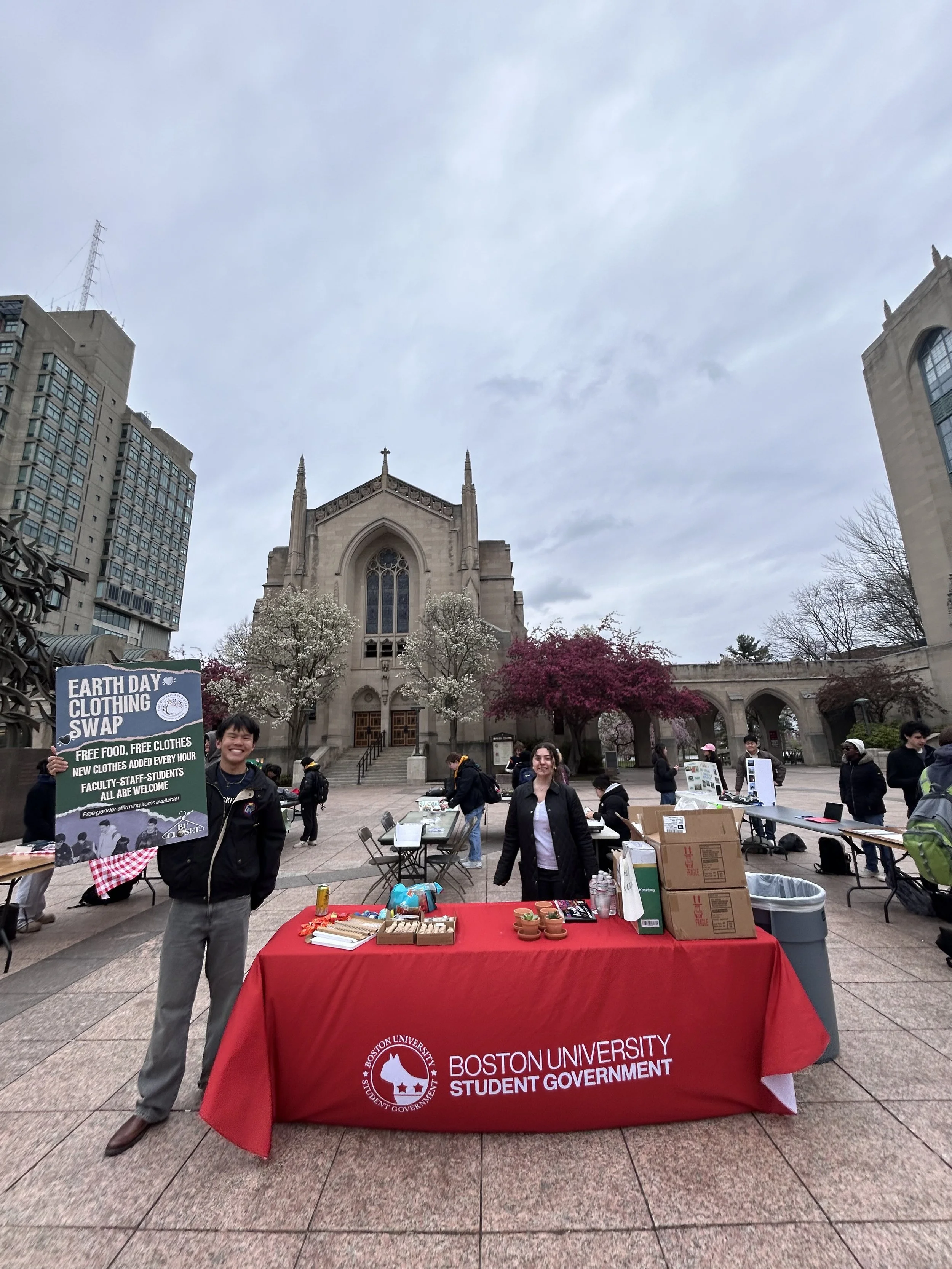 People at a Boston University Student Government event outdoors in a city plaza, with a church and tall buildings in the background. A woman is holding a sign advertising an Earth Day clothing swap. The table has a red tablecloth with the university's name and logo, and various items including snacks, boxes, and informational materials.