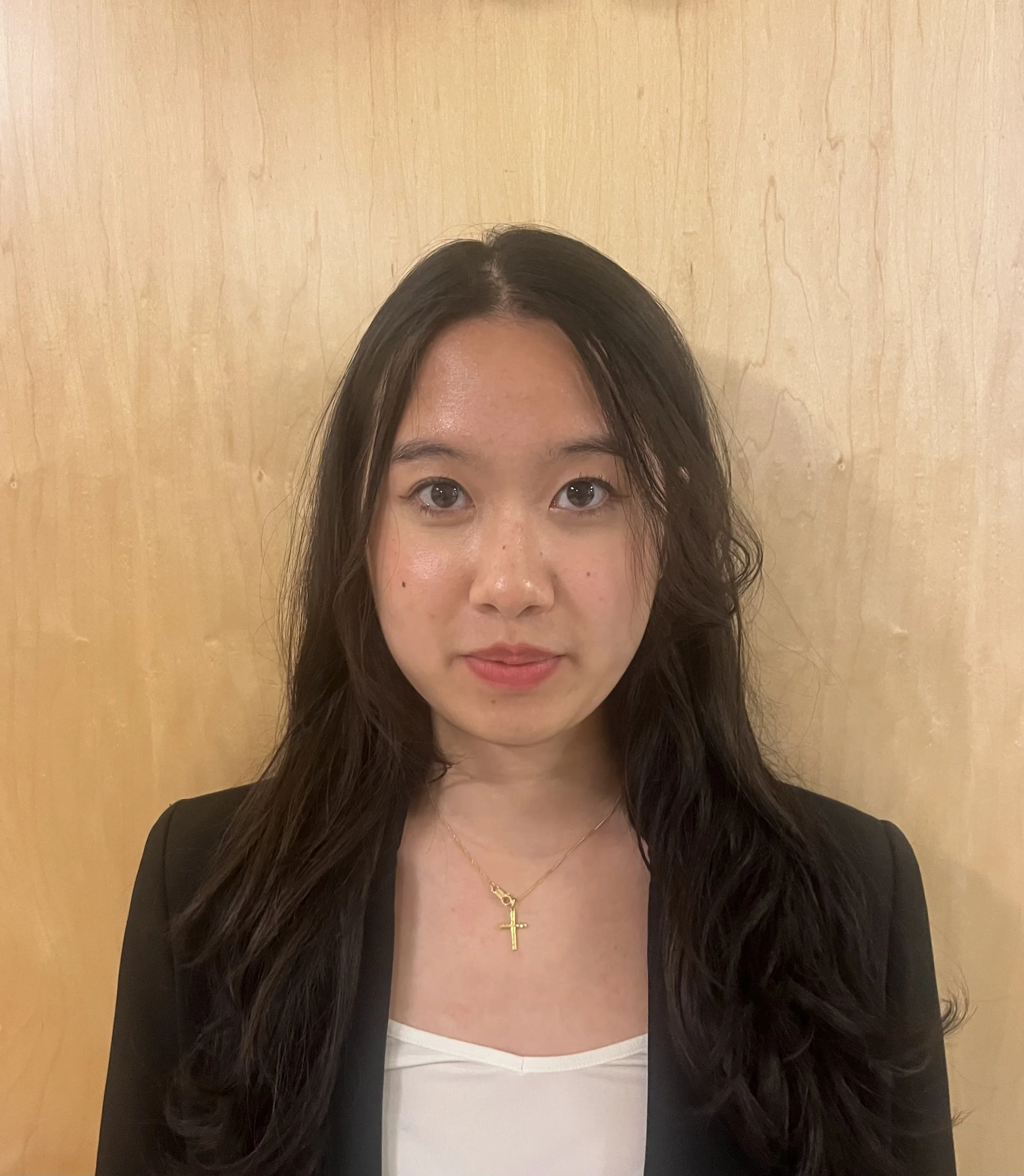 A woman with long dark hair, wearing a black blazer, a white top, a gold cross necklace, and light makeup, standing in front of a light wood-colored wall.
