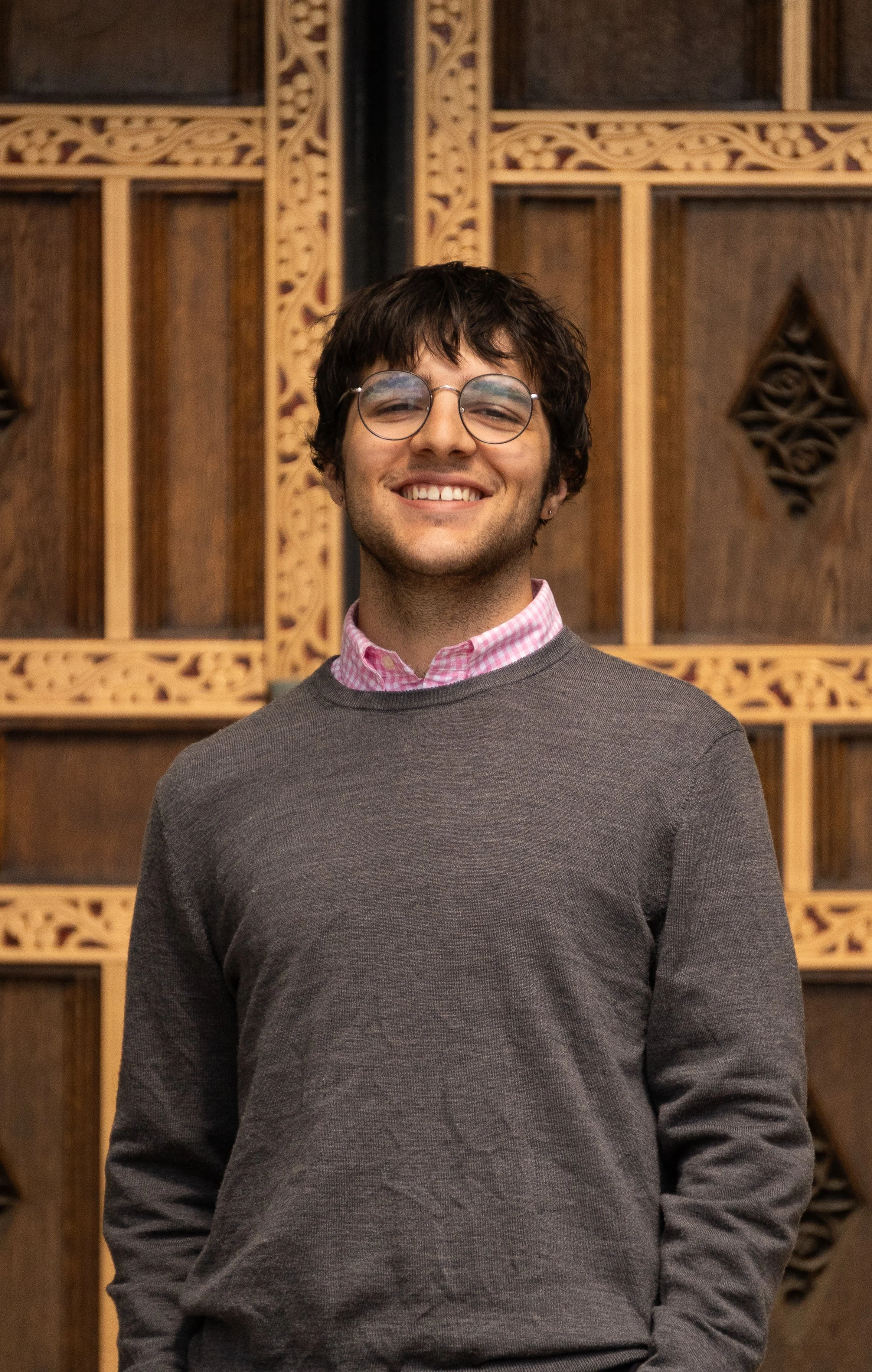 A young man with glasses smiling, wearing a pink checkered shirt under a gray sweater, standing in front of a decorative wooden wall.
